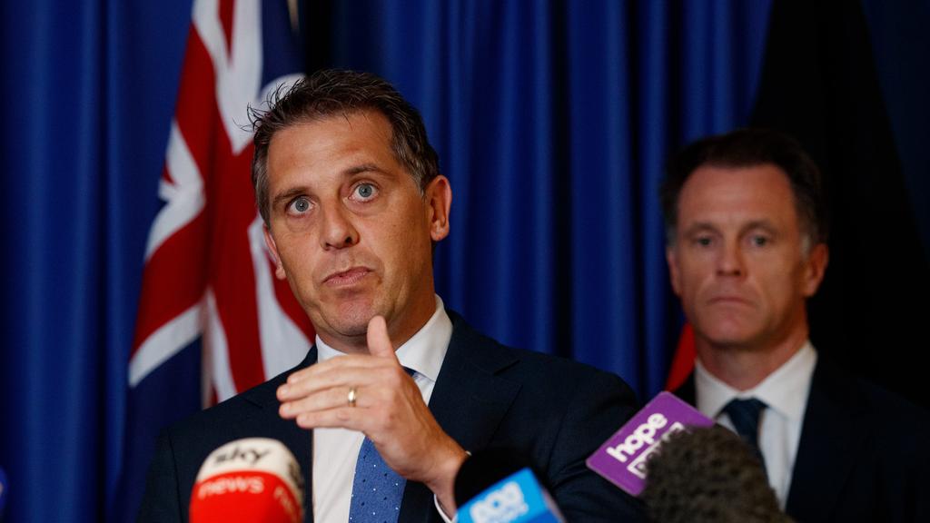 Alt text: A suited man speaks at a press conference, gesturing with his hand while standing in front of several microphones labeled with news logos like Sky News and Hope. An Australian flag is visible in the background, along with another man standing behind him, also wearing a suit.