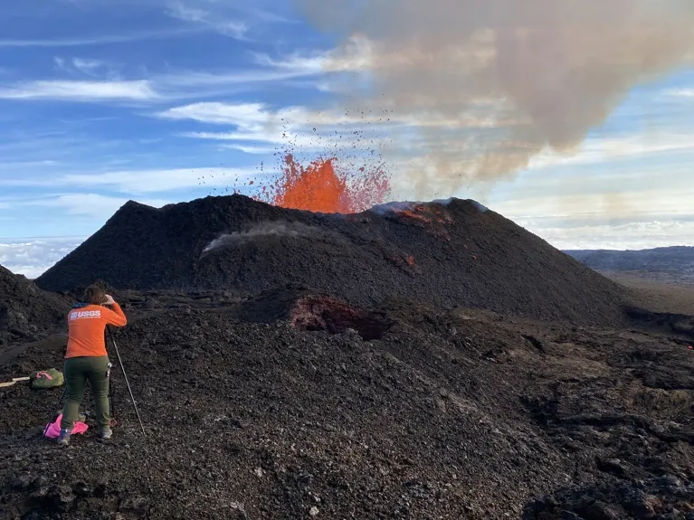 A USGS scientist in an orange shirt stands on a rocky slope, using measuring equipment to observe an active volcanic eruption in the distance. Bright orange lava shoots into the air from a dark volcanic cone, while smoke rises into a partly cloudy sky.