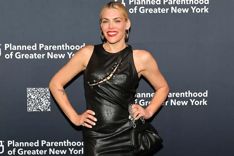 Alt text: A woman poses confidently on the red carpet at a Planned Parenthood of Greater New York event, wearing a sleeveless black leather dress with safety pin accents and holding a black handbag. The backdrop features repeating white text and a QR code.